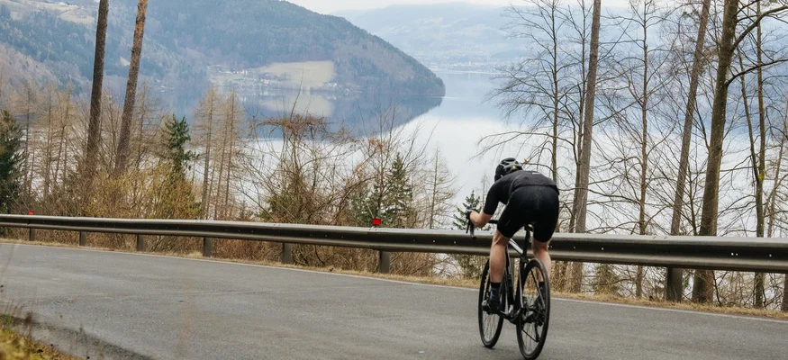 Radfahrer fährt bergab auf asphaltierter Straße mit See und Bergen im Hintergrund