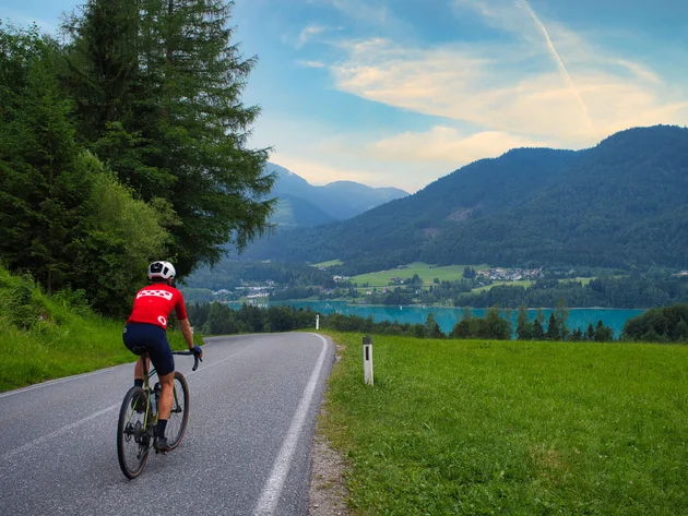 Drei Seen, eine Gravelrunde: Die 3-Seen-Tour ab Fuschl © Fuschlsee Tourismus - GravGrav Radfahrer auf Bergstraße mit See und Bergen im Hintergrund