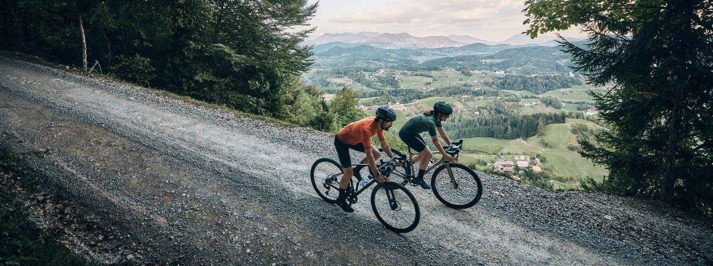 Logar Tal und Solčava Panoramastrasse © Tobias Köhler Zwei Radfahrer fahren einen schmalen Waldweg mit Bergblick hinunter