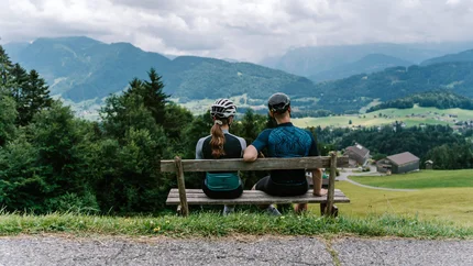 Zwei Radfahrer sitzen auf einer Bank und blicken auf die Berge und das Tal