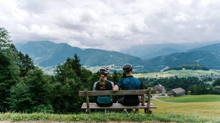 Gravelbiken in Vorarlberg: Die 7 schönsten Highlights zwischen Bodensee und Alpen © Bregenzerwald Tourismus - Christina Neubauer Zwei Radfahrer sitzen auf einer Bank und blicken auf die Berge und das Tal