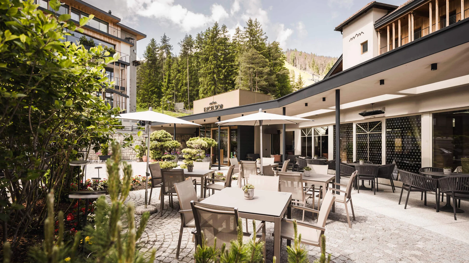 Herbst auf Schotter im Excelsior Dolomites Life Resort****s © Hannes Niederkofler Außenbereich eines Hotels mit Tischen, Stühlen und Sonnenschirmen vor Waldkulisse