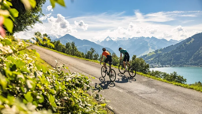 Zwei Radfahrer fahren auf Bergstraße mit See und Bergen im Hintergrund