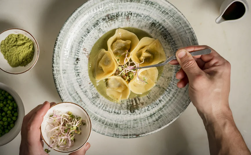 Hand plating ravioli with sprouts, surrounded by green ingredients