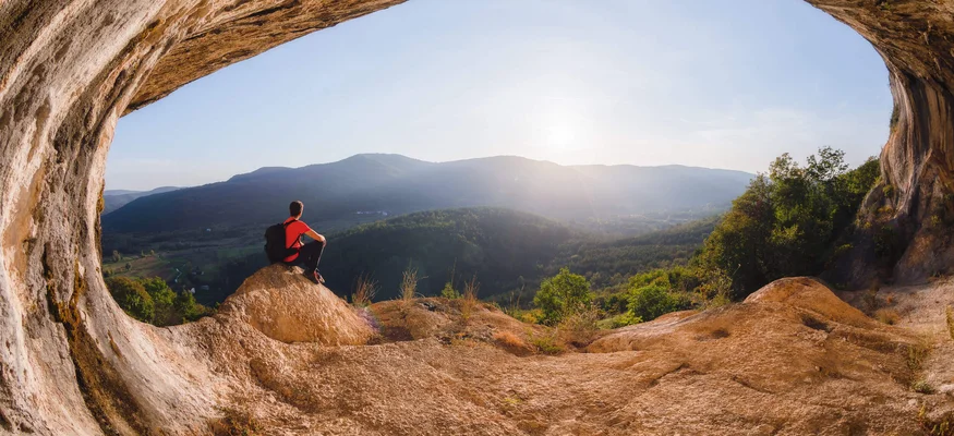 Person sitting in a cave looking out over mountains and valley at sunrise