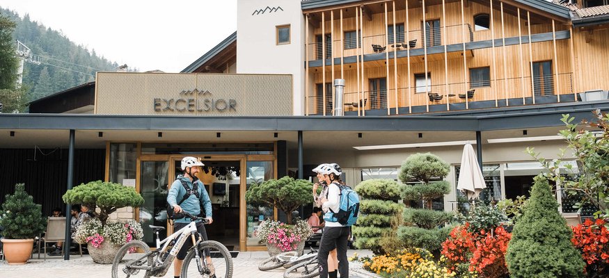 Zwei Radfahrer mit Helmen vor dem Hotel Excelsior mit blühendem Garten