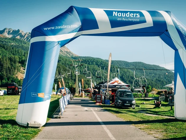 Das waren die Golden Days in Nauders © TVB-Tiroler-Oberland-Nauders-Christian-Penning - Expo GOLDEN DAYS 2026 Blau-weißer Nauders-Bogen über einer Straße vor bewaldeten Bergen und Sesselliften