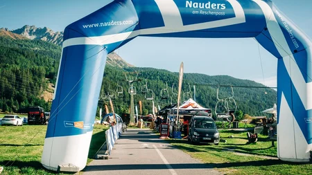 Das waren die Golden Days in Nauders © TVB-Tiroler-Oberland-Nauders-Christian-Penning - Expo GOLDEN DAYS 2026 Blau-weißer Nauders-Bogen über einer Straße vor bewaldeten Bergen und Sesselliften