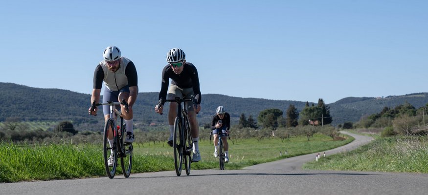 Drei Radfahrer fahren auf einer Landstraße in ländlicher Gegend bei klarem Himmel.