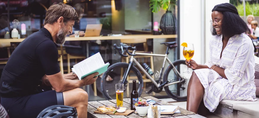 Müde Beine, volle Bäuche, offene Herzen © Stefan Leitner Mann liest Buch, Frau trinkt Getränk an Caféterrasse mit Fahrrad im Hintergrund