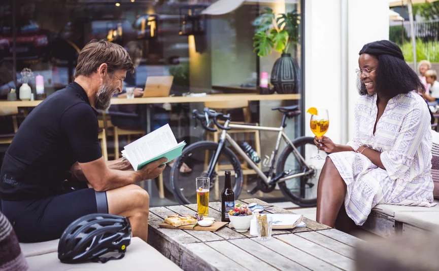 Mann liest Buch, Frau trinkt Getränk an Caféterrasse mit Fahrrad im Hintergrund