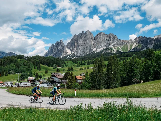 Zwei Radfahrer fahren auf einer Straße mit Alpenbergen und grüner Landschaft im Hintergrund