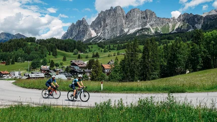 Two cyclists riding on a road with alpine mountains and green landscape in the background