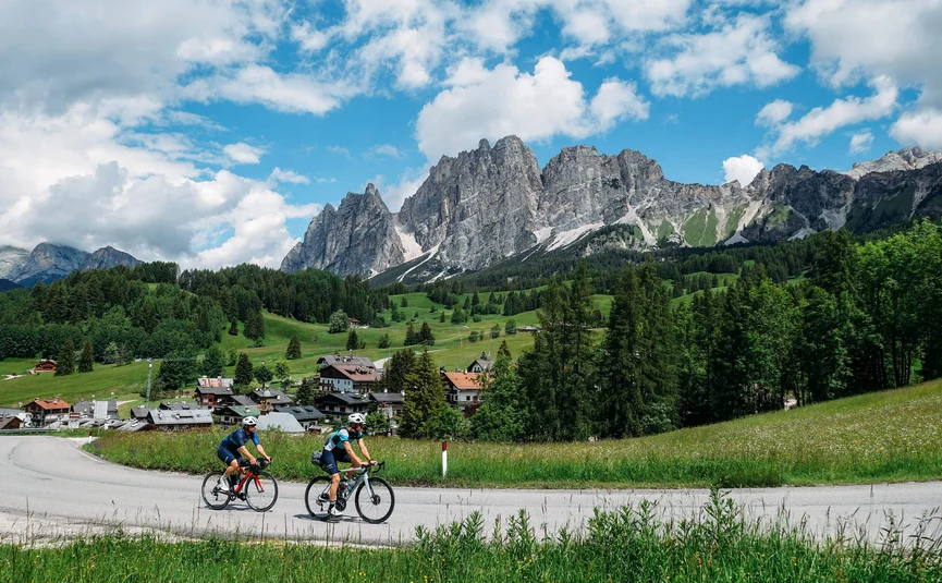Zwei Radfahrer fahren auf einer Straße mit Alpenbergen und grüner Landschaft im Hintergrund