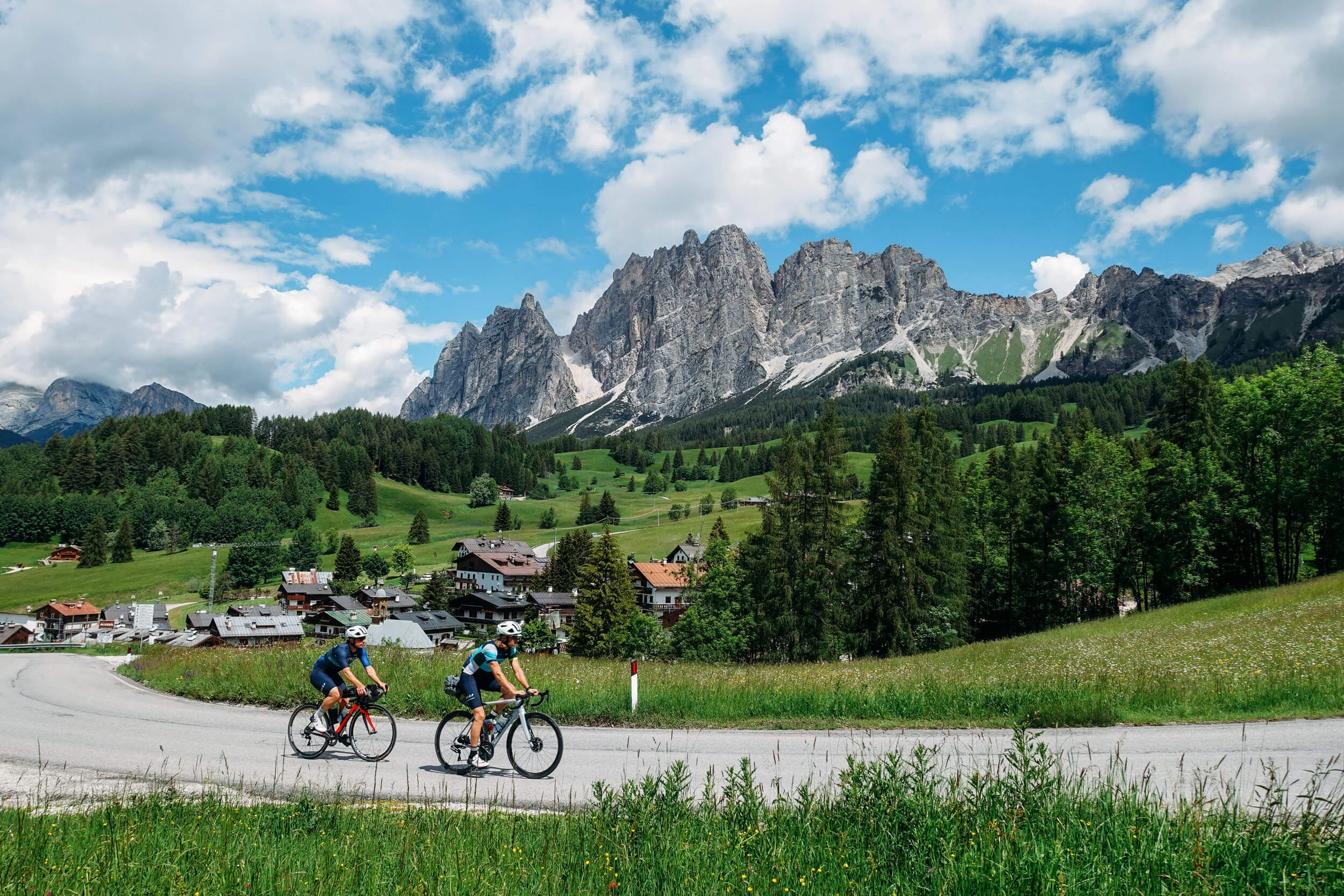 Zwei Radfahrer fahren auf einer Straße mit Alpenbergen und grüner Landschaft im Hintergrund