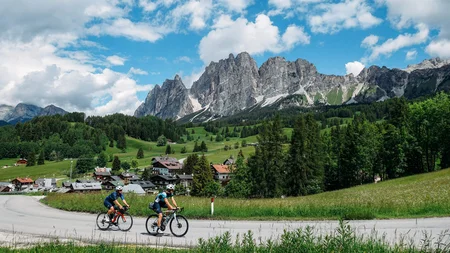 Zwei Radfahrer fahren auf einer Straße mit Alpenbergen und grüner Landschaft im Hintergrund