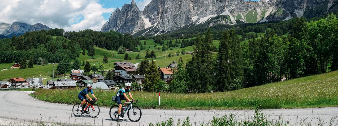 Cinque Torri © Hotel Melodia del Bosco Two cyclists riding on a road with alpine mountains and green landscape in the background