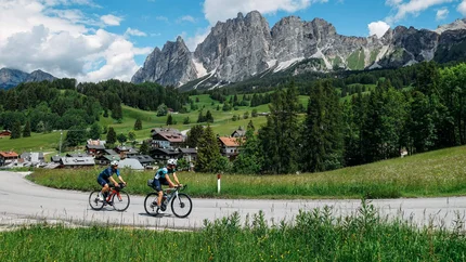 Zwei Radfahrer fahren auf einer Straße mit Alpenbergen und grüner Landschaft im Hintergrund