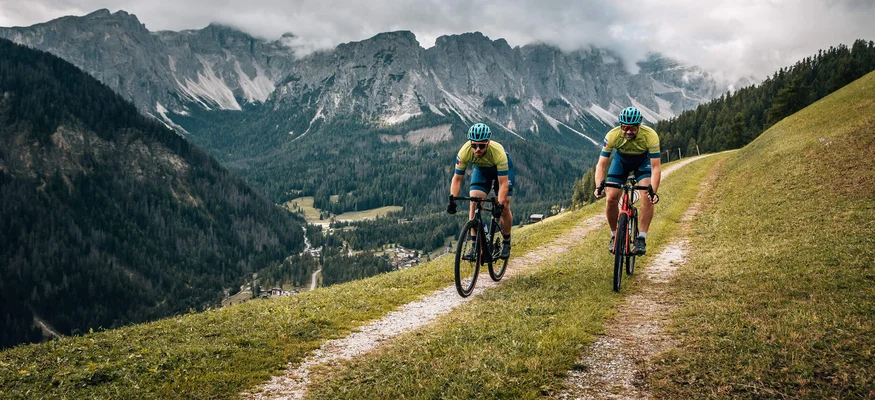 Zwei Radfahrer fahren auf einem Bergweg mit Alpen im Hintergrund
