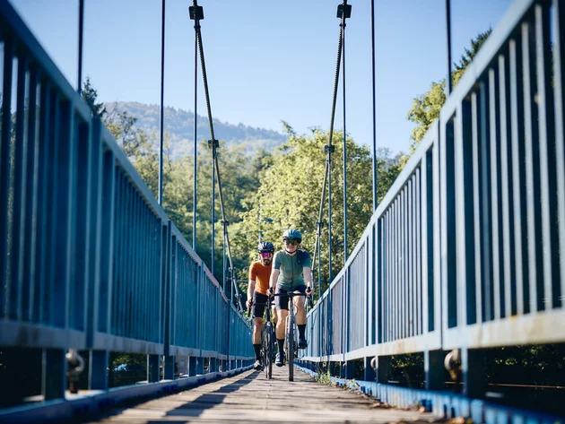 Two cyclists riding on a suspension bridge surrounded by greenery and clear sky