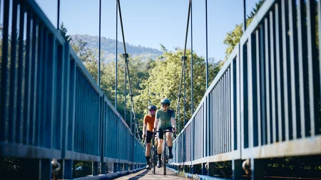Two cyclists riding on a suspension bridge surrounded by greenery and clear sky