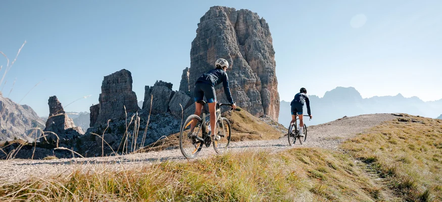 Zwei Radfahrer fahren auf einem Bergweg vor großer Felsformation im Sonnenschein