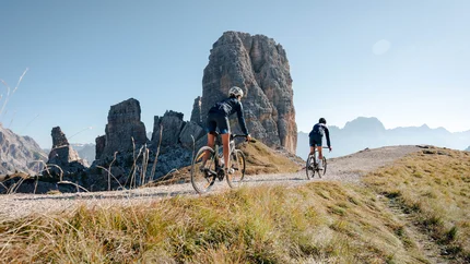 Two cyclists riding on mountain trail with large rock formation in sunny weather