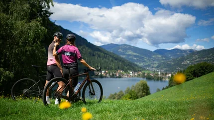Zwei Radfahrer mit Helm stehen auf einer Wiese mit Blick auf See und Berge