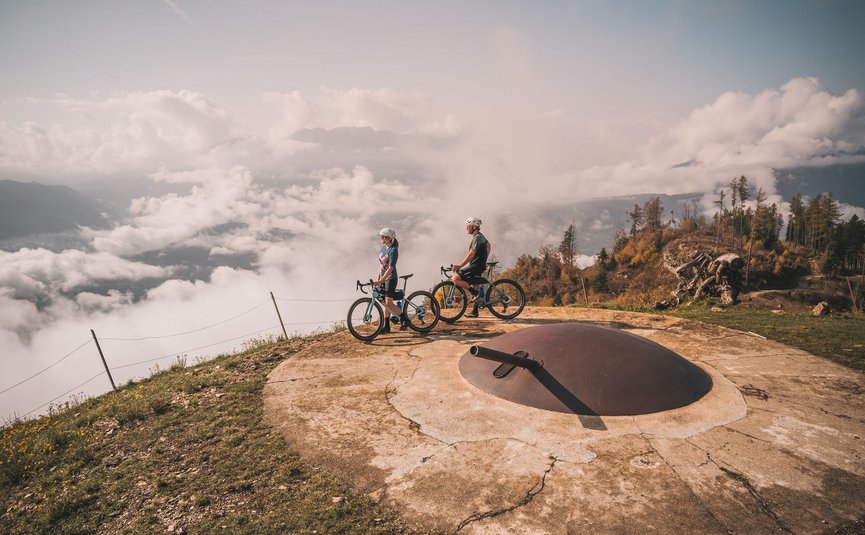 Two cyclists on mountain near old military bunker with fog in valley