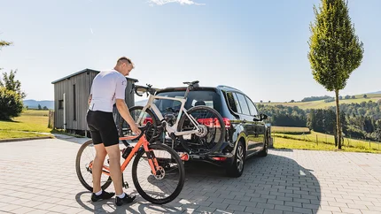 Neue Gravelbike Holidays Hotels 2026 © Stefan J. Wolf Photography Mann befestigt Fahrrad an Fahrradträger hinten am Auto auf sonnigem Landparkplatz