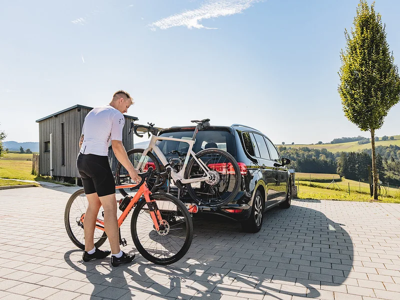 Gravelbike Holidays © Stefan J. Wolf Photography Mann befestigt Fahrrad an Fahrradträger hinten am Auto auf sonnigem Landparkplatz
