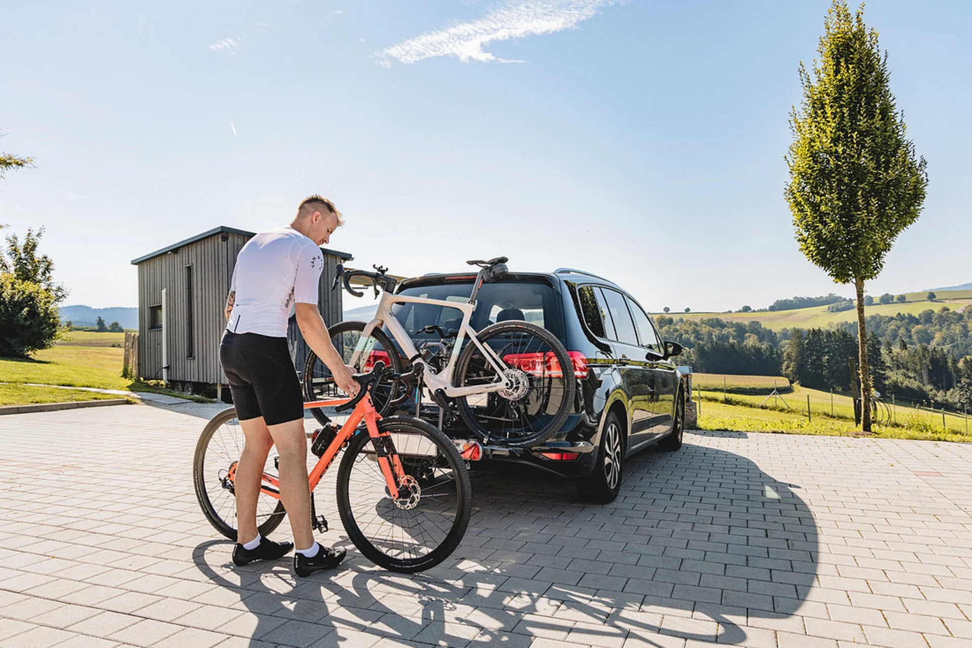 Gravelbike Holidays © Stefan J. Wolf Photography Mann befestigt Fahrrad an Fahrradträger hinten am Auto auf sonnigem Landparkplatz