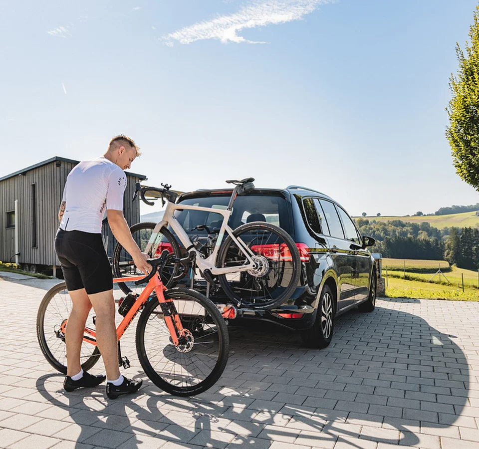 Neue Gravelbike Holidays Hotels 2026 © Stefan J. Wolf Photography Mann befestigt Fahrrad an Fahrradträger hinten am Auto auf sonnigem Landparkplatz