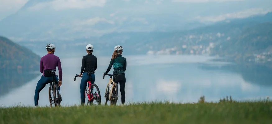 NOCKBIKE Area © Martin Hofmann Drei Radfahrer mit Helmen blicken auf einen See und Berge