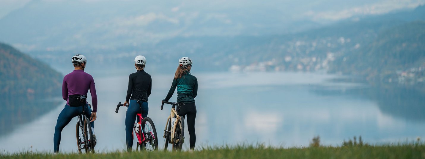 Der Mittelpunkt Kärntens © Martin Hofmann Drei Radfahrer mit Helmen blicken auf einen See und Berge