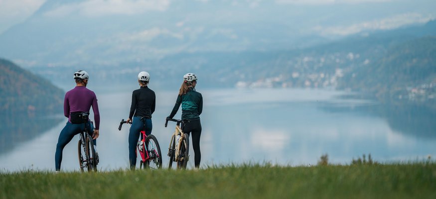 Drei Radfahrer mit Helmen blicken auf einen See und Berge