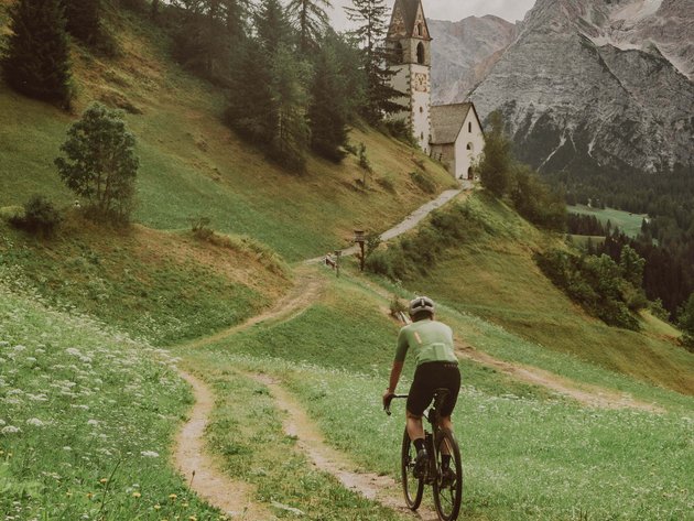 Alpine Gravel Experience © John Braynard Radfahrer fährt auf einem Weg mit Kirche und Bergen im Hintergrund