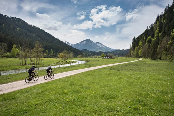 Österreich © Arjan Kruik FreeWheeler Zwei Radfahrer auf einem Weg in einer grünen Berglandschaft mit Fluss und Haus