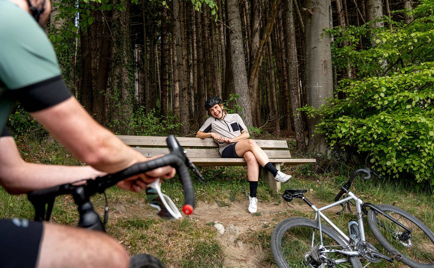 Frau mit Fahrradhelm sitzt lachend auf einer Bank im Wald, Mann auf Fahrrad im Vordergrund