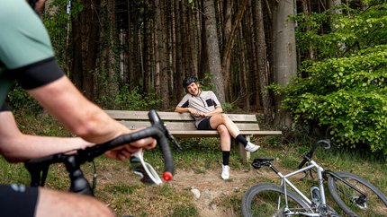 Graveltour Feldkirch © Chris Gollhofer - Vorarlberg Tourismus Frau mit Fahrradhelm sitzt lachend auf einer Bank im Wald, Mann auf Fahrrad im Vordergrund