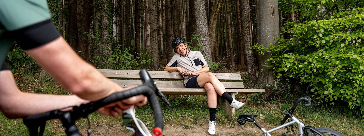Graveltour Feldkirch © Chris Gollhofer - Vorarlberg Tourismus Frau mit Fahrradhelm sitzt lachend auf einer Bank im Wald, Mann auf Fahrrad im Vordergrund