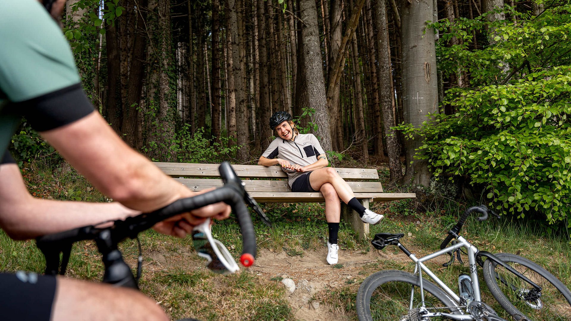Frau mit Fahrradhelm sitzt lachend auf einer Bank im Wald, Mann auf Fahrrad im Vordergrund