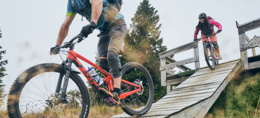 Zwei Mountainbiker fahren eine Rampe auf einem Waldweg hinunter
