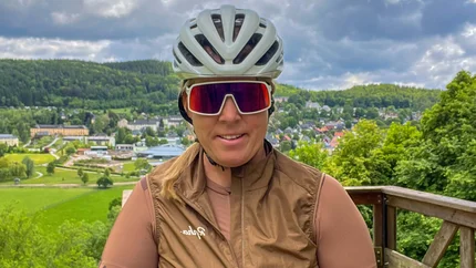 Frau mit Fahrradhelm und Sportbrille auf Aussichtsplattform mit Wald und Ort im Hintergrund