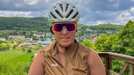 Frau mit Fahrradhelm und Sportbrille auf Aussichtsplattform mit Wald und Ort im Hintergrund