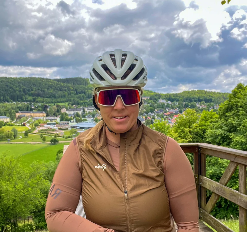 Frau mit Fahrradhelm und Sportbrille auf Aussichtsplattform mit Wald und Ort im Hintergrund