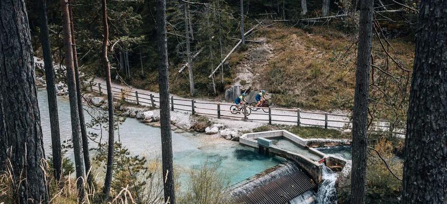 Zwei Radfahrer auf einem Waldweg neben einem klaren Bach mit kleinem Wehr