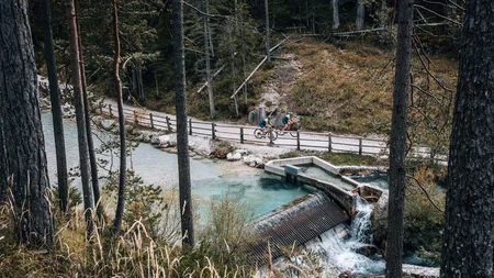 Two cyclists on forest path beside clear stream with small dam