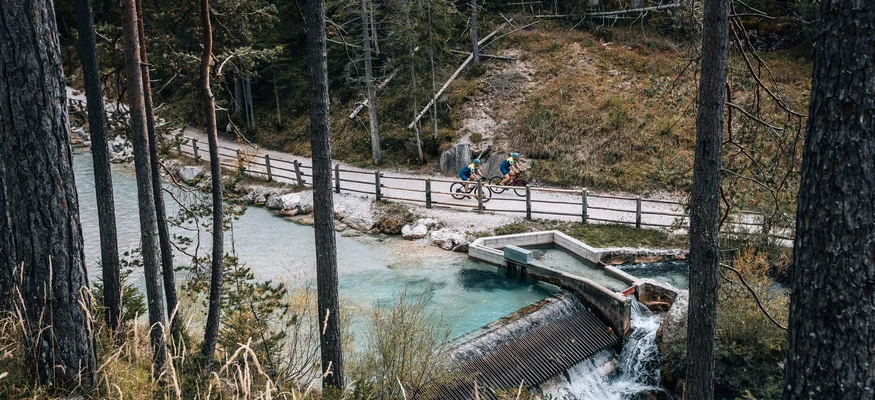 Zwei Radfahrer auf einem Waldweg neben einem klaren Bach mit kleinem Wehr
