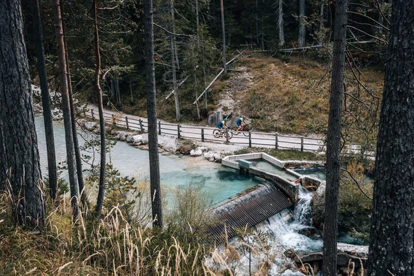 Italien © Jens Scheibe Zwei Radfahrer auf einem Waldweg neben einem klaren Bach mit kleinem Wehr